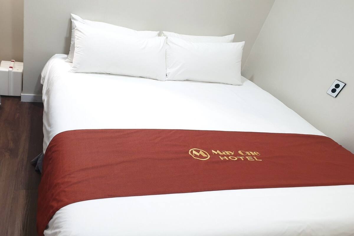 A hotel room with a bed, white bedding, a red blanket with a logo, a small bedside table, and a wall socket.