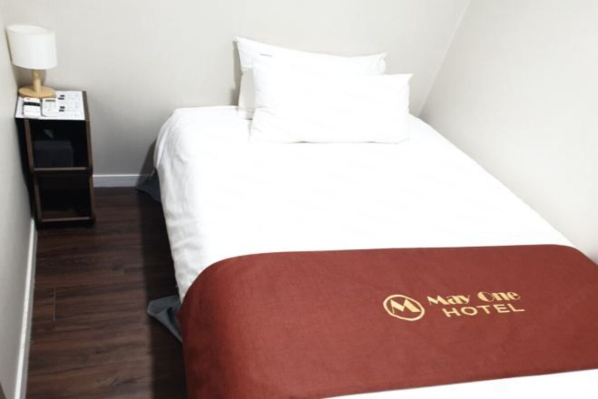 A hotel room with a bed, white bedding, a brown throw blanket with the hotel logo, a nightstand with a lamp, and a wooden floor.