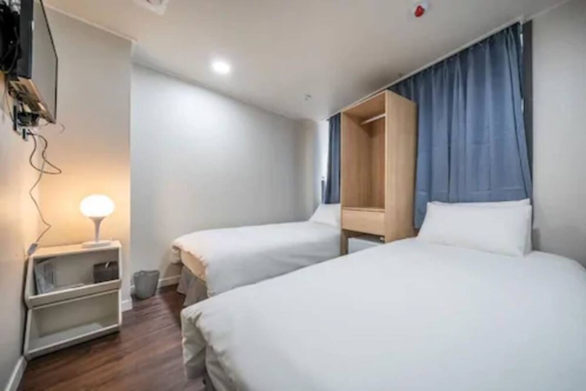 A hotel room with two beds, a wardrobe, a bedside table with a lamp, and a TV mounted on the wall.