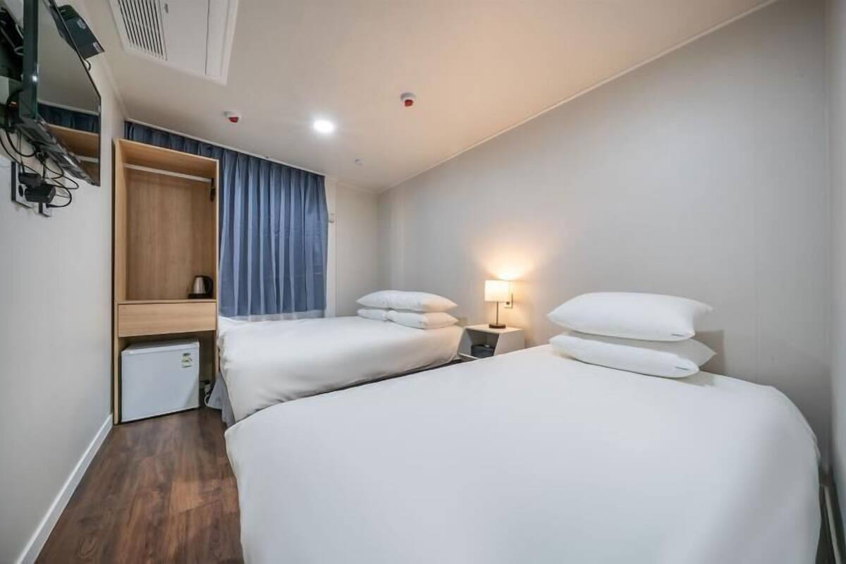 A hotel room with two beds, a small desk, a wardrobe, and a ceiling-mounted air conditioning unit.