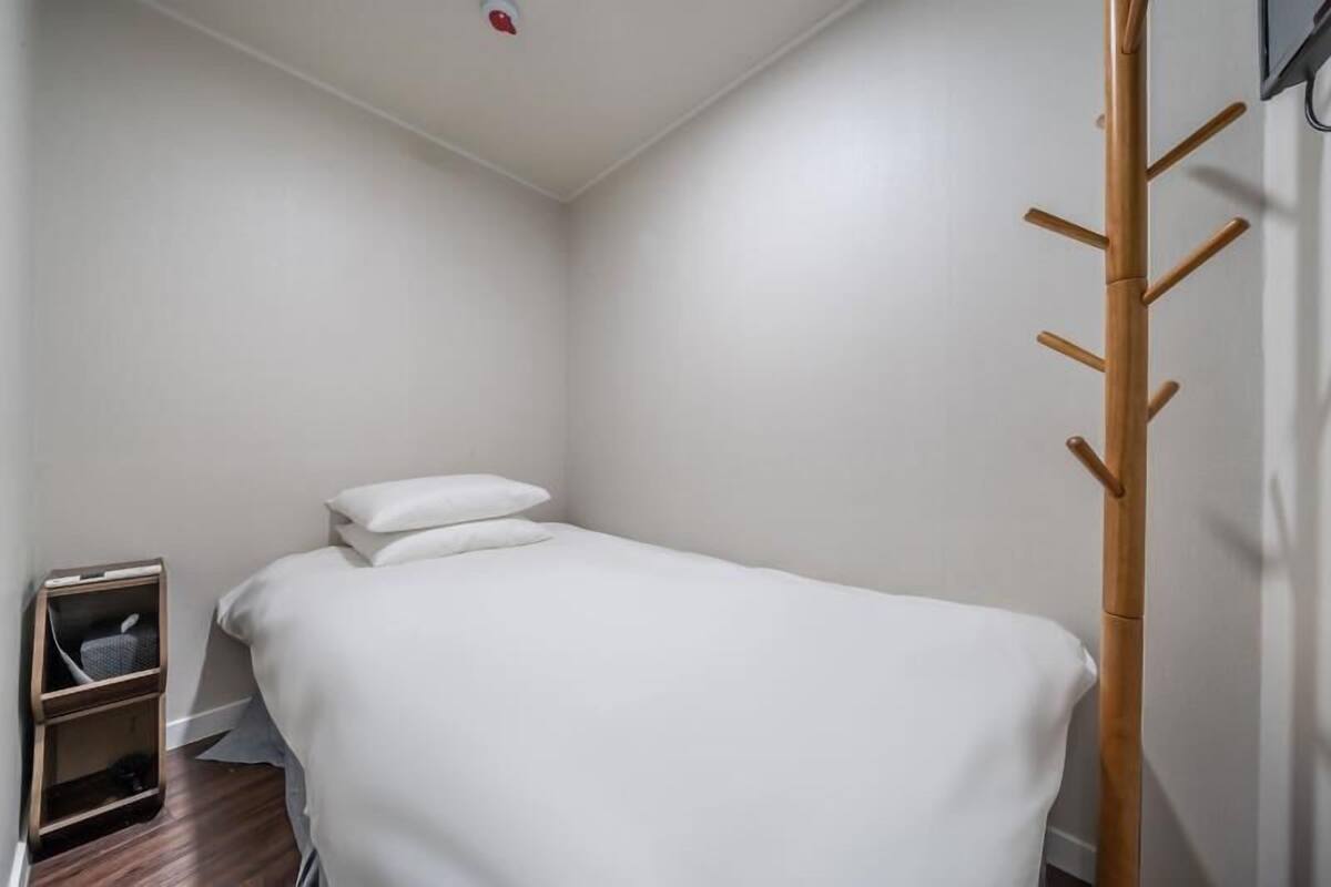 A small, neatly made room with a single bed, a wooden coat rack, and a small shelf unit.