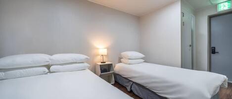 A hotel room with two single beds, a nightstand with a lamp, a door with an emergency exit sign, and a wooden floor.