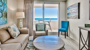 A room with a beige sofa, a round wooden table, a blue chair, a lamp, and a view of the beach through a large window.