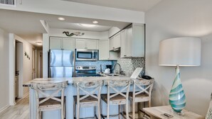 A kitchen with white cabinets, a stainless steel refrigerator, a microwave, a stove, and a bar area with wooden stools.