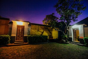 A nighttime view of a residential property with a well-lit entrance, a small garden, and a tree casting a shadow on the wall.