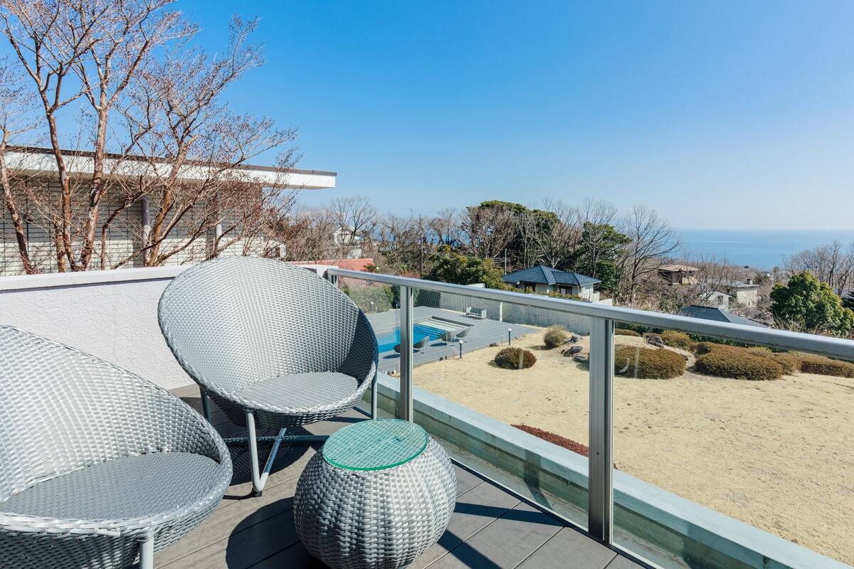 A balcony with wicker furniture, a glass railing, and a view of the sea and distant buildings.