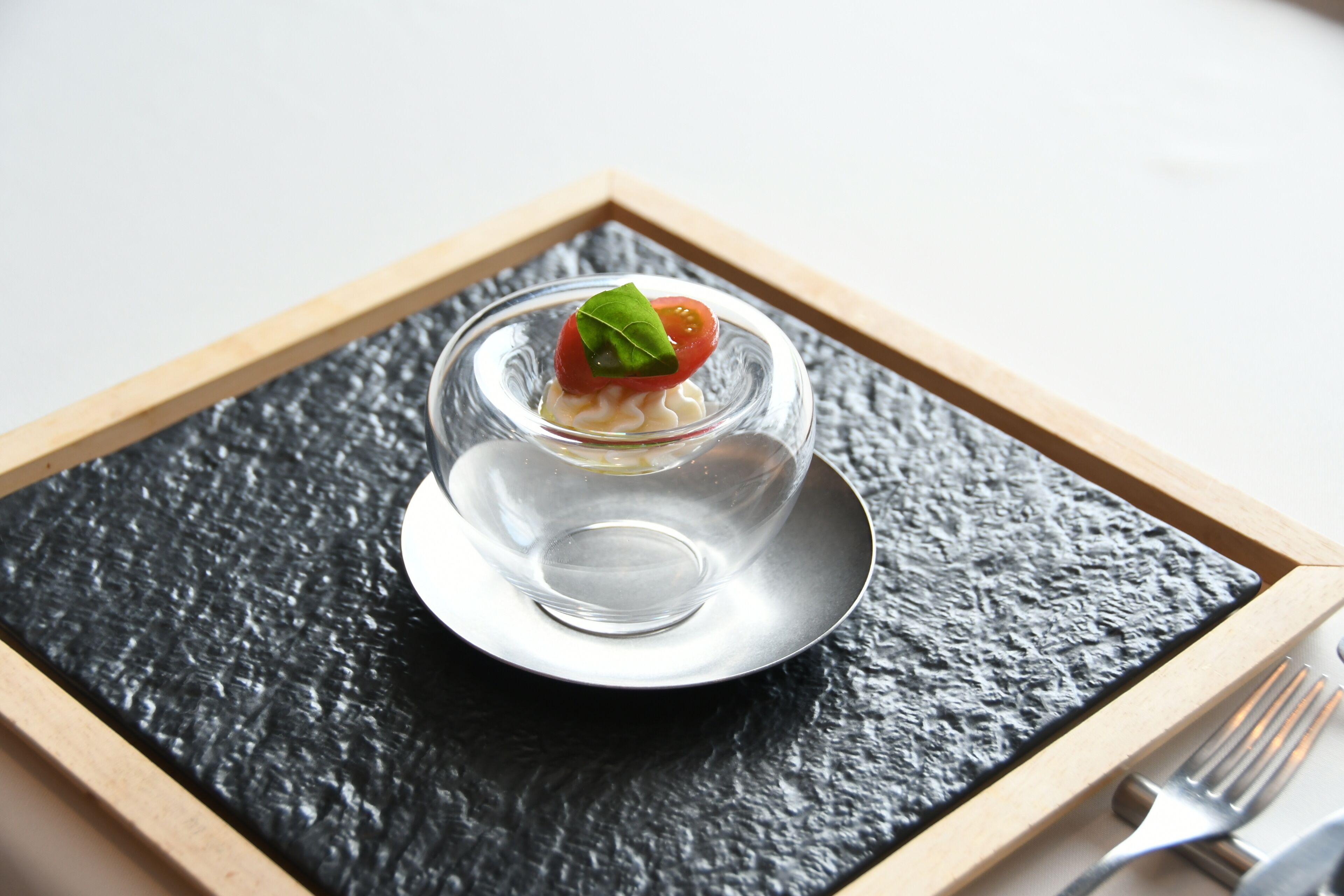 A clear glass dessert on a black slate plate, framed by a wooden border, with a fork beside it.