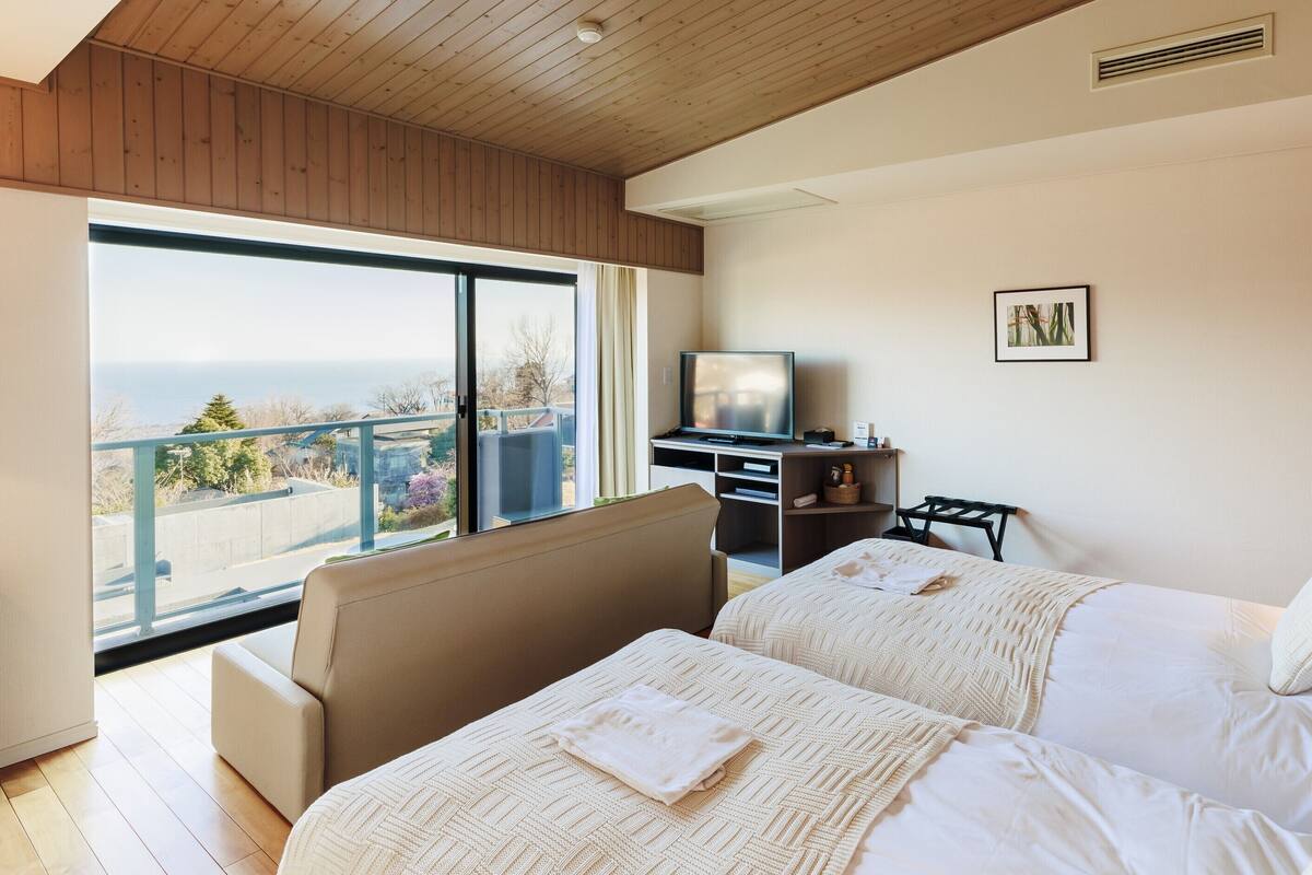 A hotel room with two beds, a television, a desk, and a large window offering a view of the sea.