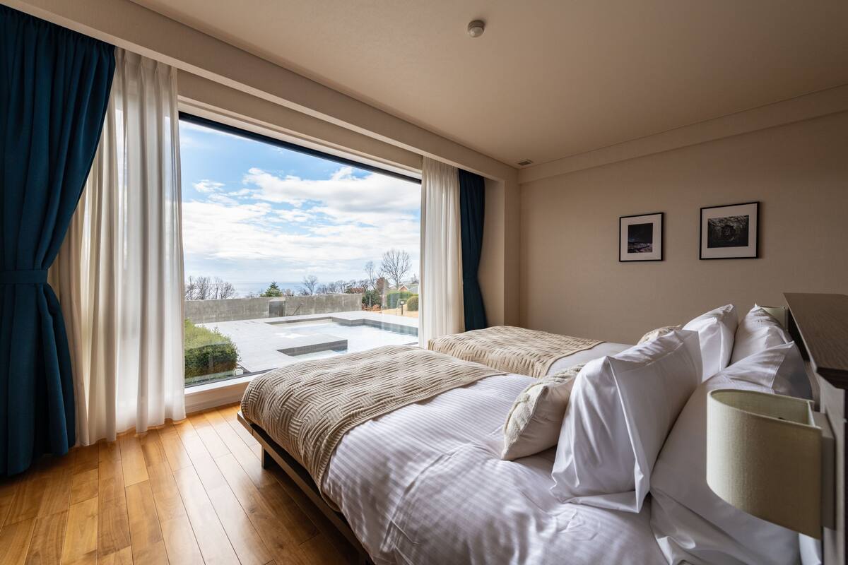 A bedroom with two beds, a large window, and a view of a rooftop pool.