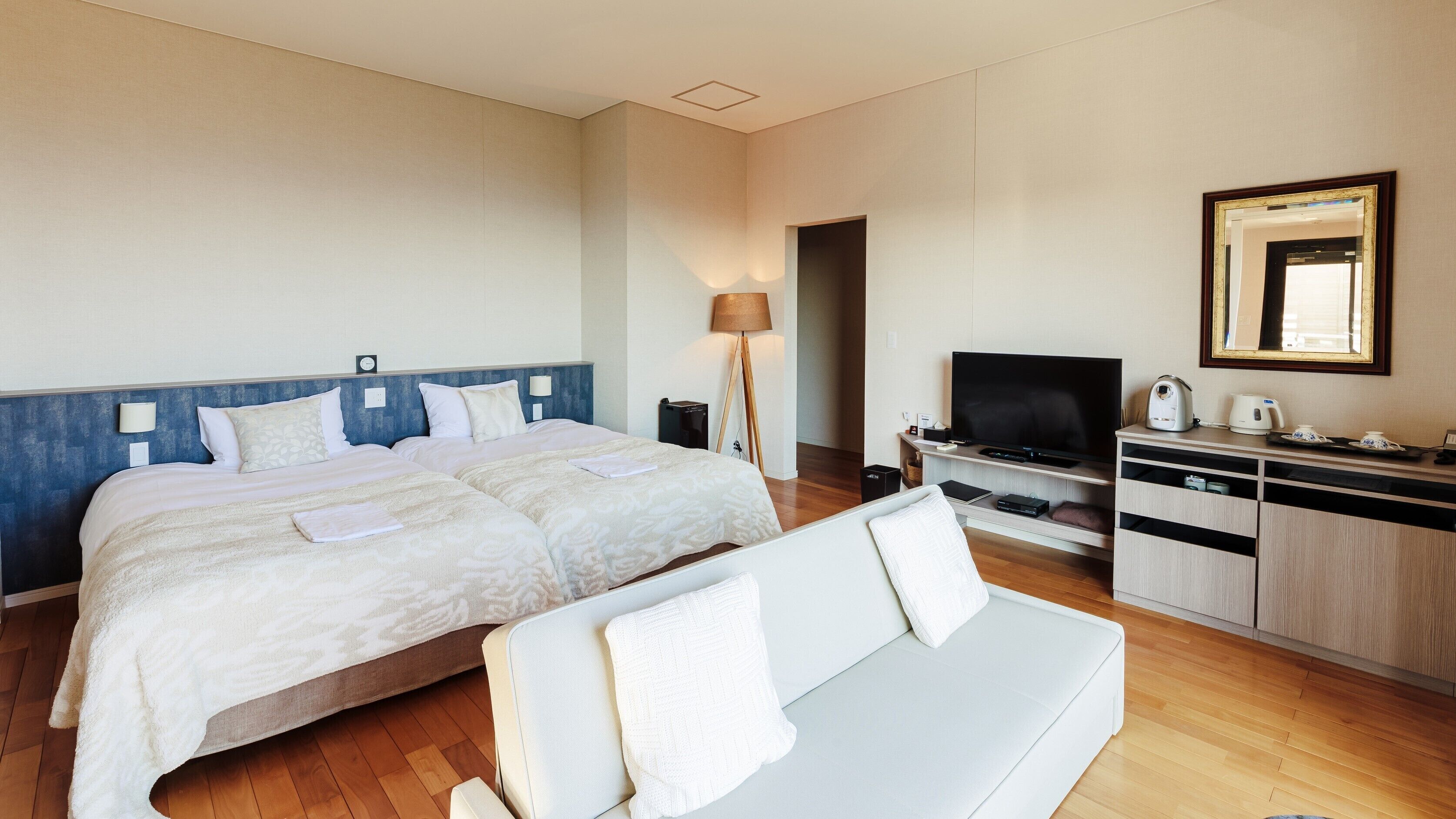 Ein Hotelzimmer mit zwei Betten, einem Fernseher, einem Schreibtisch mit Wasserkocher und einer Couch.