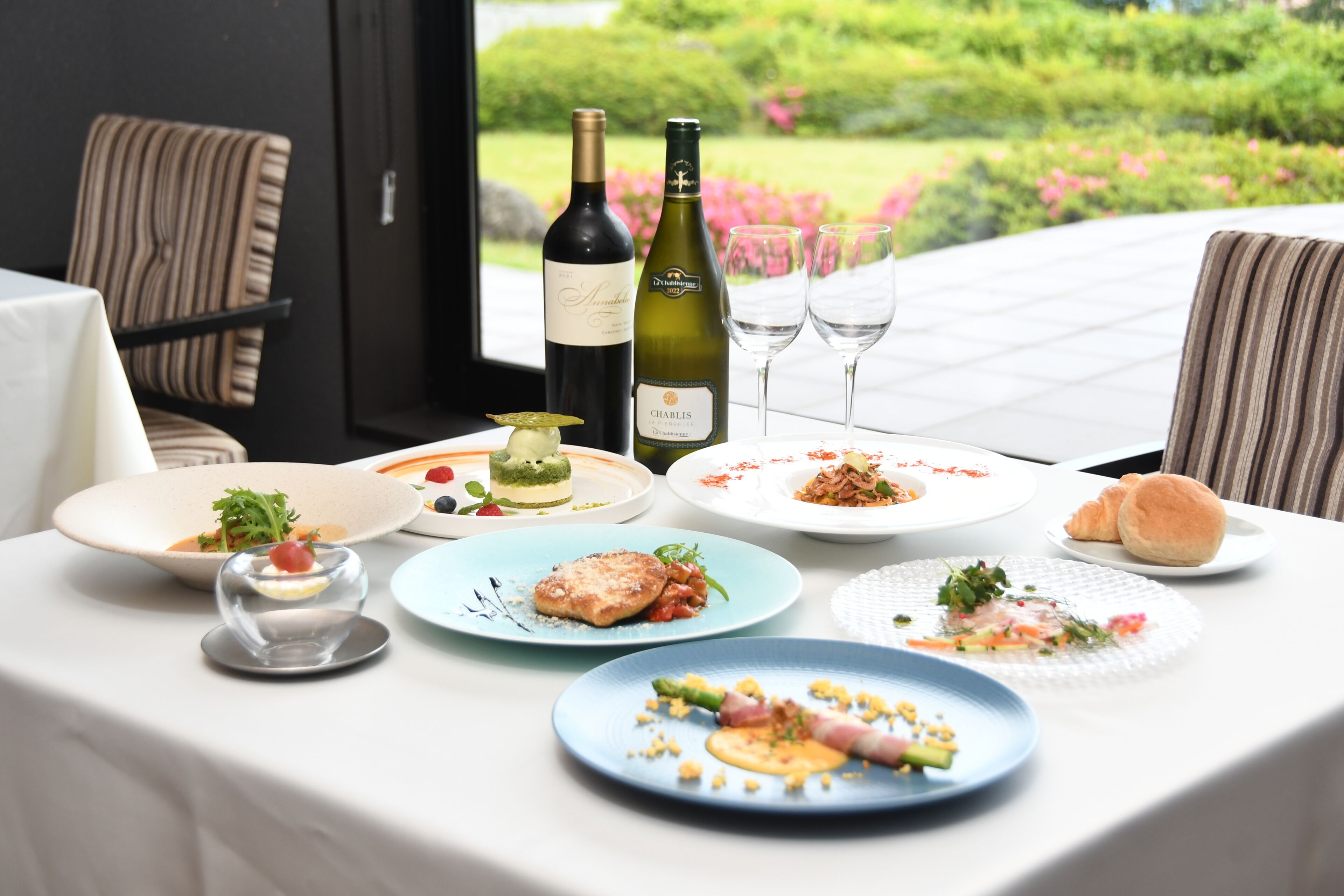 A table set with plates of food, two wine bottles, and glasses, with a view of a garden.