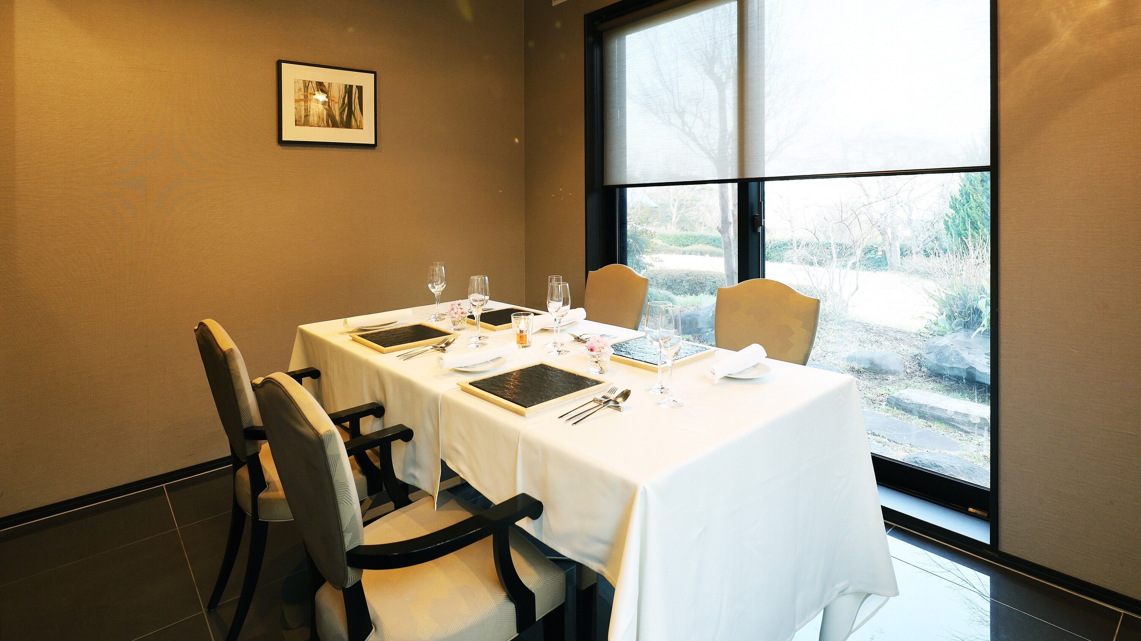 Uma sala de jantar com uma mesa posta para quatro pessoas, cadeiras e uma janela com vista para árvores.