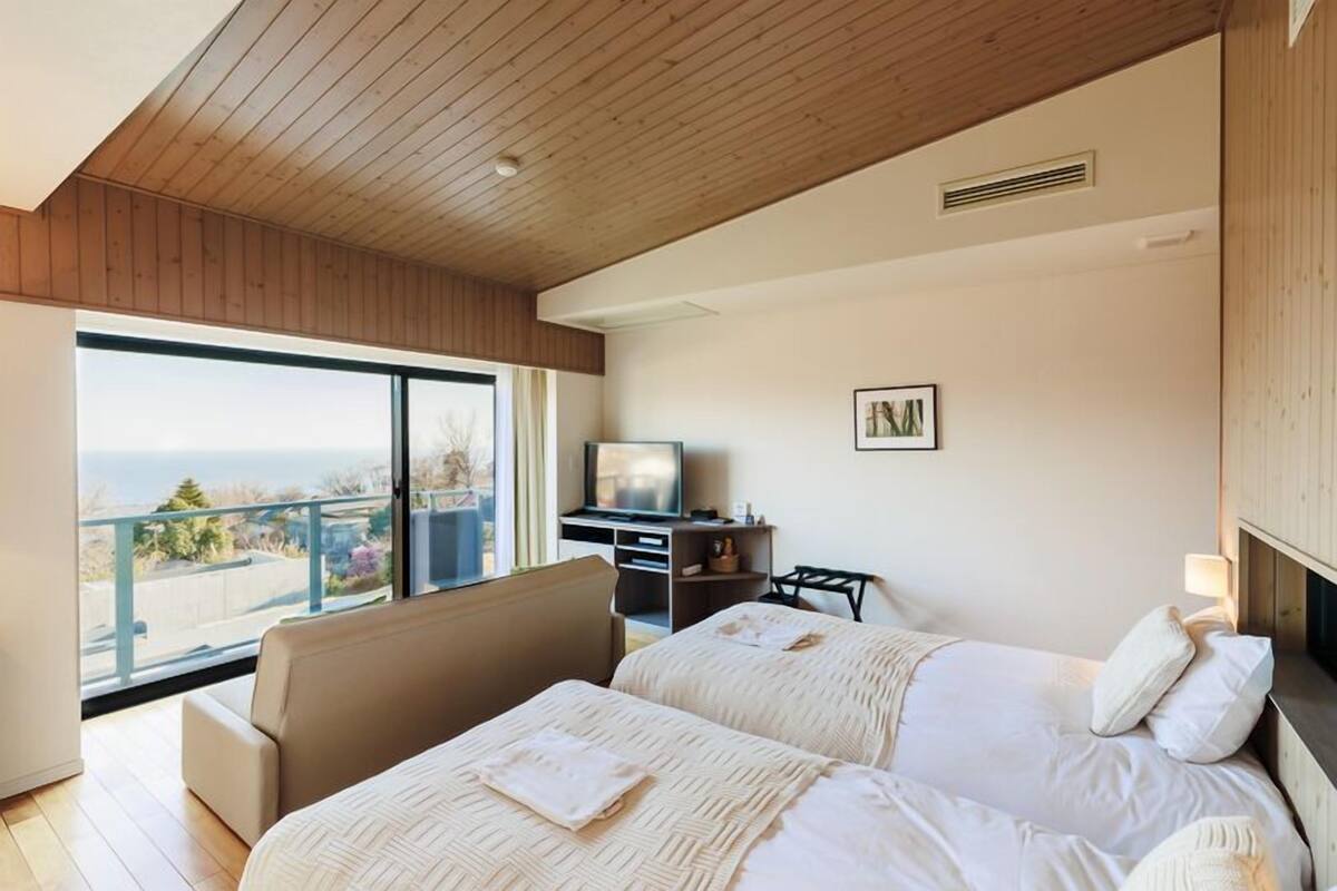 A hotel room with two beds, a TV, a desk, and a large window with a view.