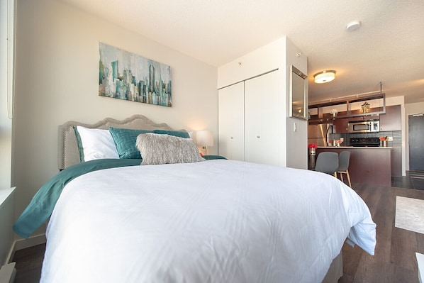 A hotel room with a large bed, a cityscape painting, a kitchenette, and a separate dining area.