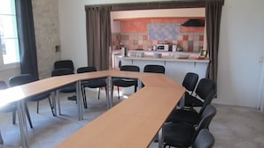 Une salle de conférence avec une longue table et des chaises, un coin cuisine équipé d’un micro-ondes et d’une machine à café, et une fenêtre avec des rideaux.