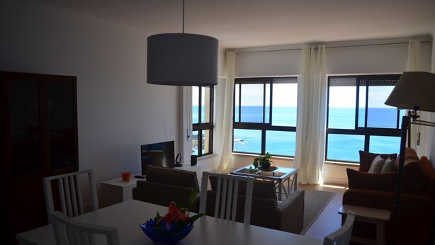 A modern living room with a dining area, large windows with ocean views, a ceiling lamp, and a small table with a plant.