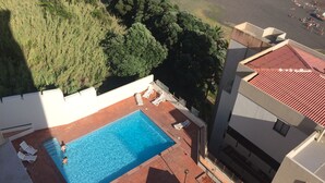 An aerial view of a rooftop swimming pool with a few people swimming, surrounded by greenery and adjacent buildings.