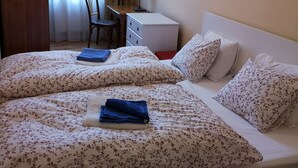 Ein ordentlich bezogenes Bett mit Blumenmuster, ein Schreibtisch mit Lampe und ein Sessel in einem Zimmer mit Fenster und Vorhängen.