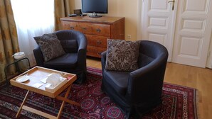 Ein Wohnzimmer mit zwei schwarzen Ledersesseln, einem Holztisch, einem Fernseher auf einer Holzkommode, einem gemusterten Teppich und einer Tür mit weißem Feld.