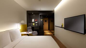 A hotel room with a bed, a desk with a chair, a TV mounted on the wall, and a small table with a lamp.