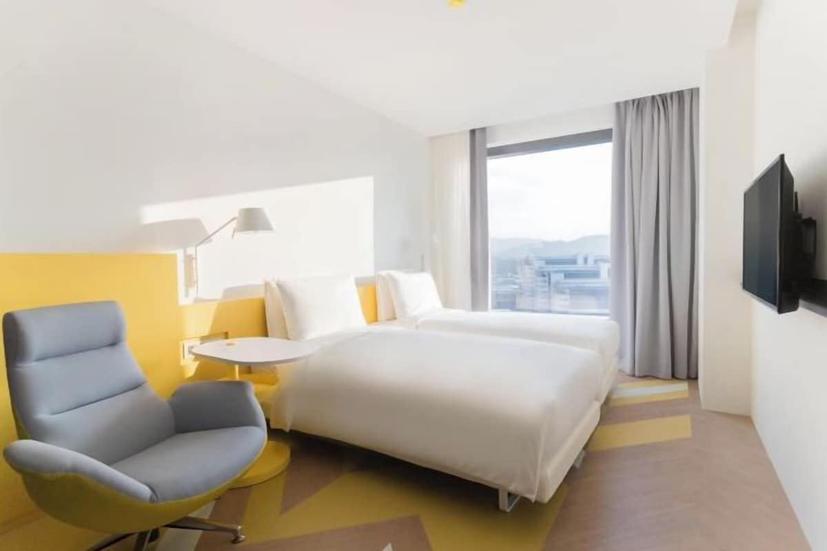 A hotel room with a bed, a chair, a small table, a TV, and a large window with curtains.