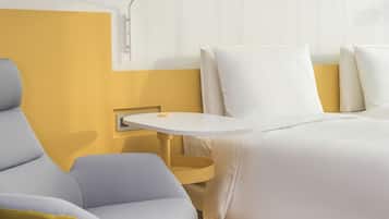 A modern hotel room with a yellow accent wall, a white bed, a white nightstand, a white lamp, and a yellow and gray chair.
