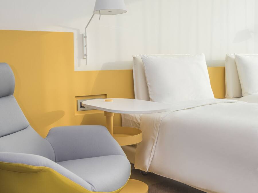 A modern hotel room with a yellow accent wall, a white bed, a white nightstand, a white lamp, and a yellow and gray chair.