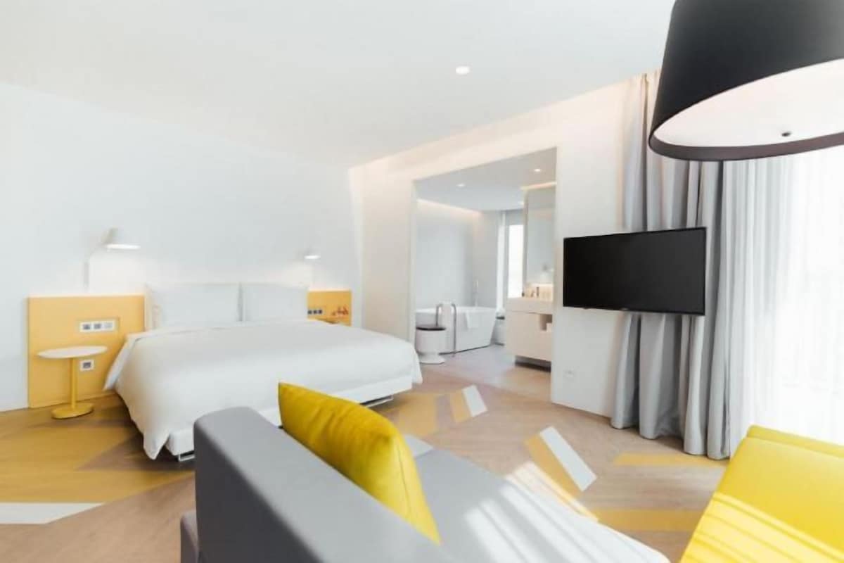 A modern hotel room with a large bed, a flat-screen TV, a sofa with yellow cushions, and a small table.