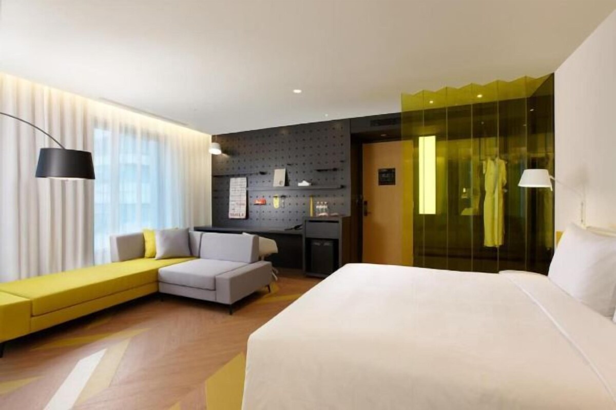 A modern hotel room with a large bed, a sofa, a desk, and a glass partition.