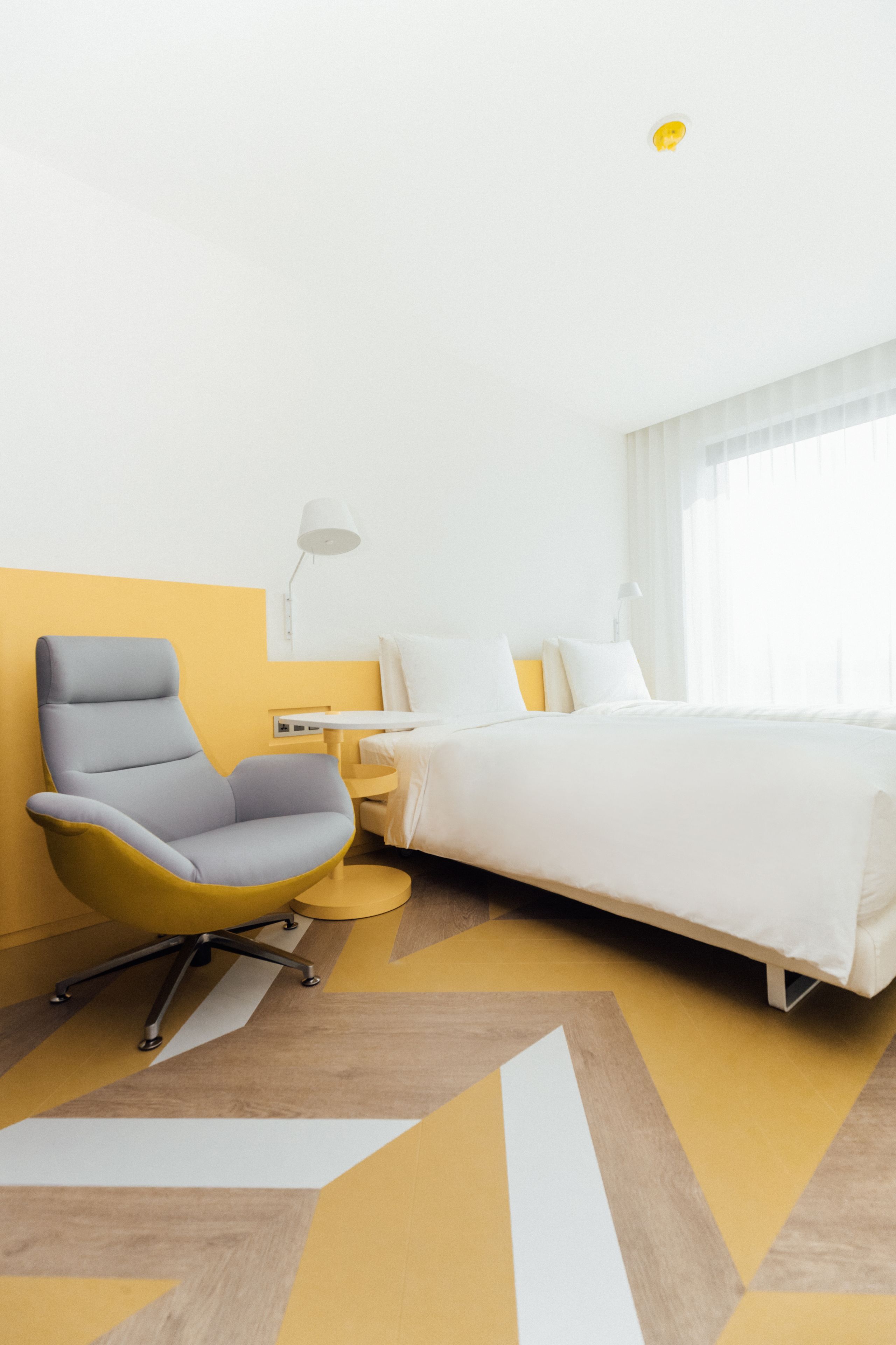 Quarto de hotel moderno com uma cama grande, uma escrivaninha com abajur e uma cadeira única em amarelo e cinza.