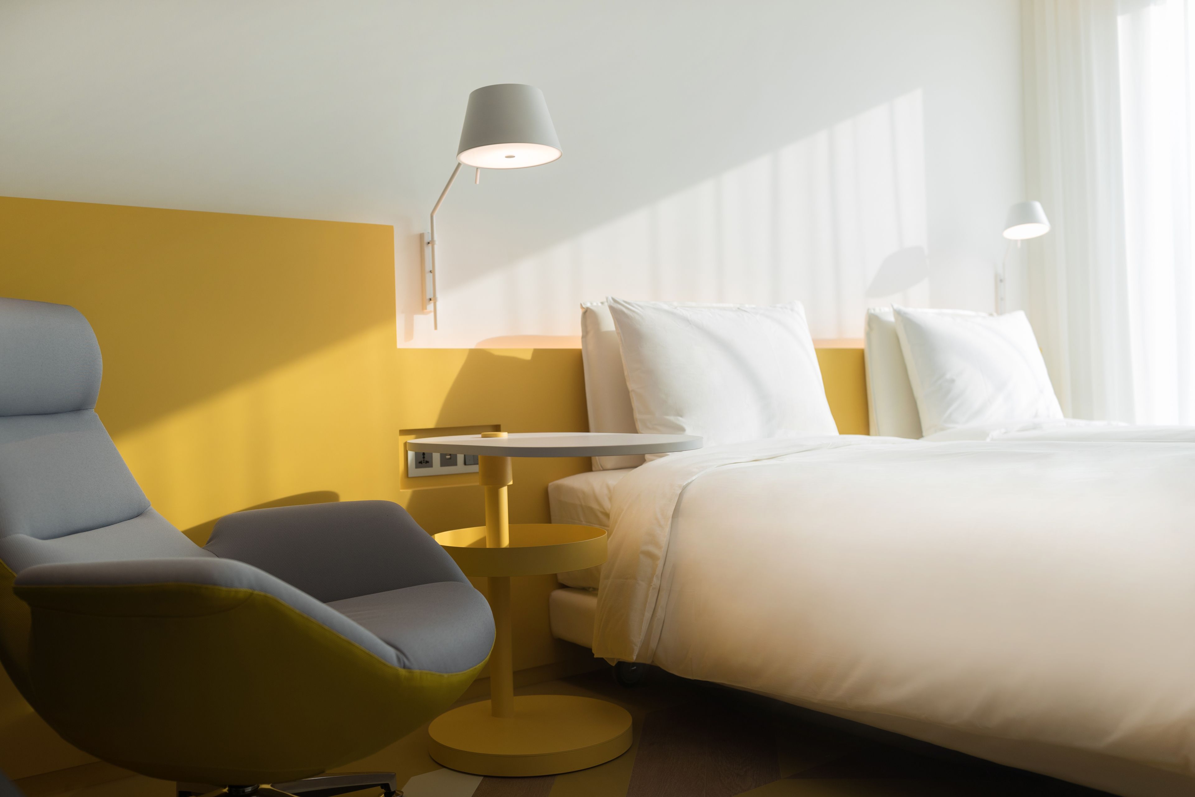 A modern hotel room with a bed, a desk, a chair, and a lamp.