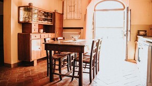 A rustic kitchen with a wooden table and chairs, a cabinet with glassware, and a window with curtains.