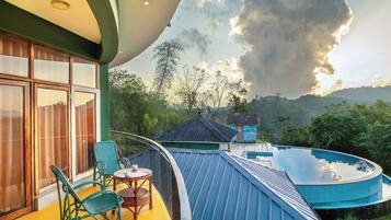A balcony with wicker chairs and a small table, overlooking a swimming pool and a traditional house.