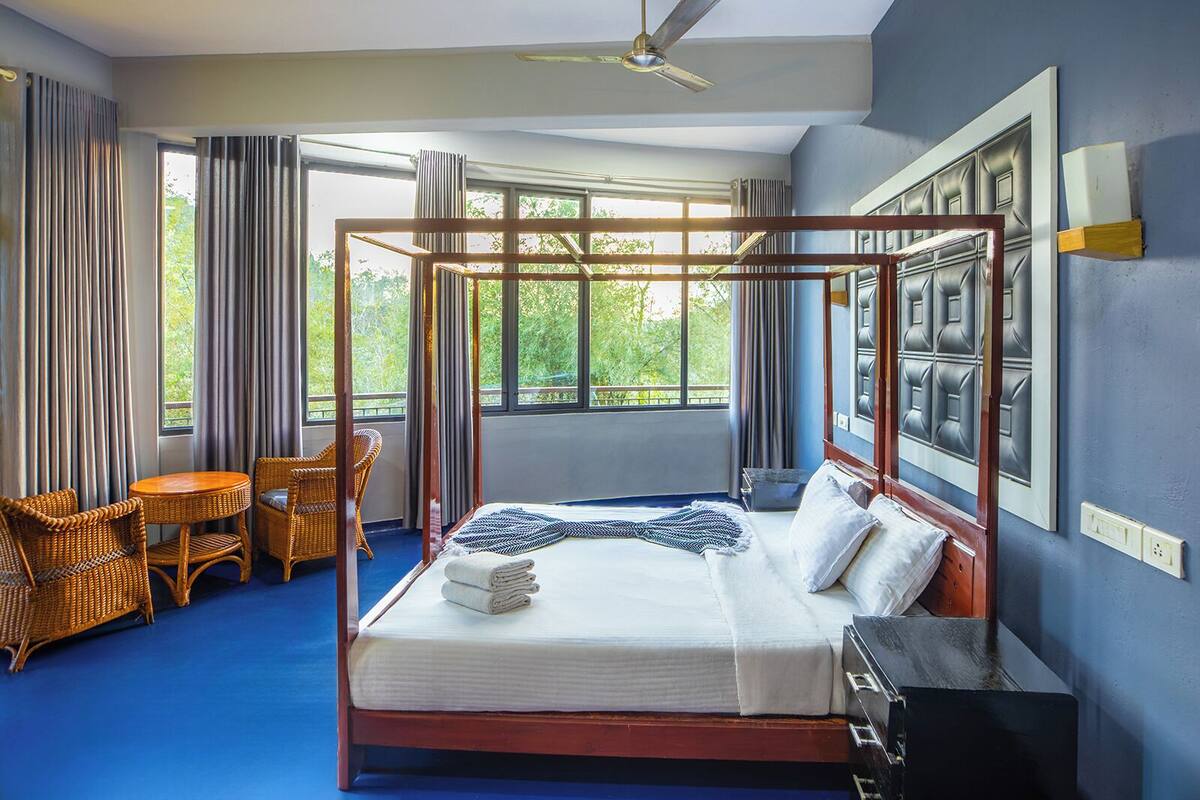 Una habitación con cama con dosel, ventilador de techo, un ventanal con cortinas, sillas de mimbre y alfombra azul.