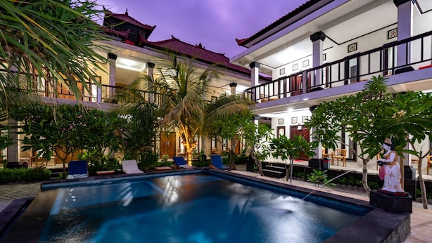 A swimming pool with lounge chairs, surrounded by tropical plants and a multi-story building with balconies. - Mega Cahya Bungalow (Lembongan Island)