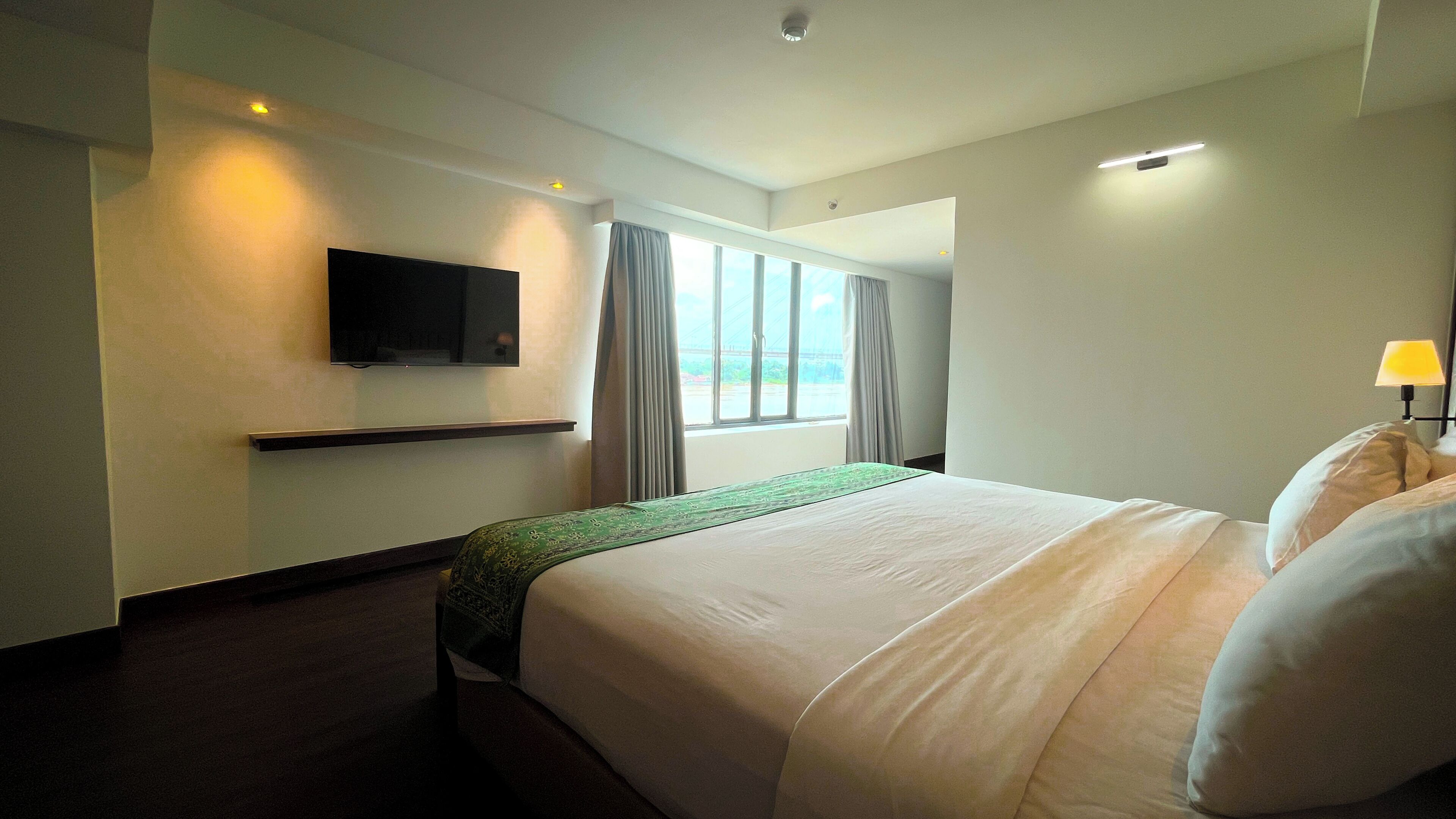 A hotel room with a bed, a flat-screen TV mounted on the wall, a window with curtains, and a bedside lamp.