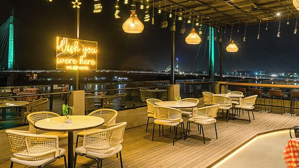 A rooftop bar with wicker chairs and tables, a neon sign, and a view of the city skyline at night. - Wiltop Hotel (Jambi)
