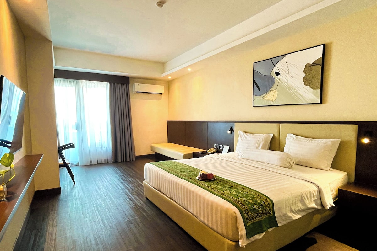 A hotel room with a large bed, a desk, a chair, a bedside table, and a painting on the wall.