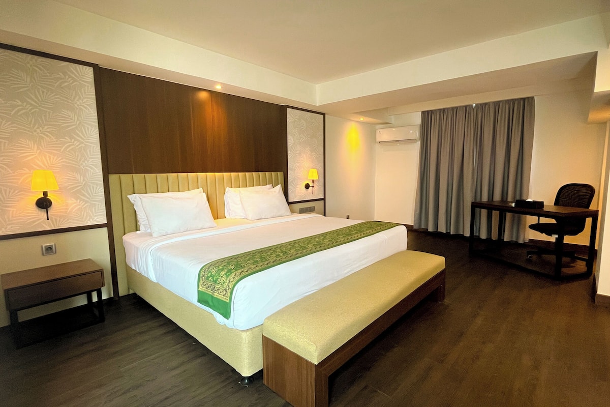 A hotel room with a bed, bedside lamps, a desk, a chair, a bench, and an air conditioning unit.