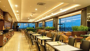 A spacious dining area with long tables and chairs, large windows with a city view, and a bar area with shelves. - Wiltop Hotel (Jambi)
