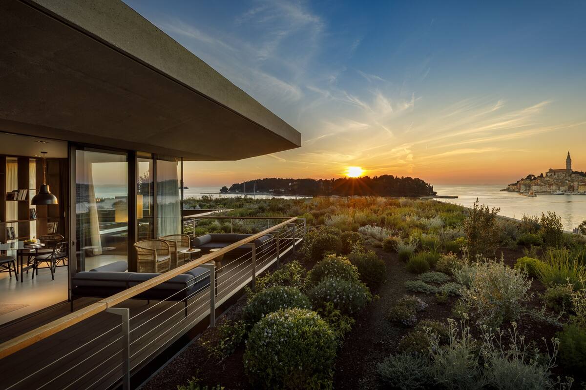 Un área de estar moderna al aire libre con vistas a la puesta de sol sobre una masa de agua, un horizonte urbano lejano y una colina con una torre.