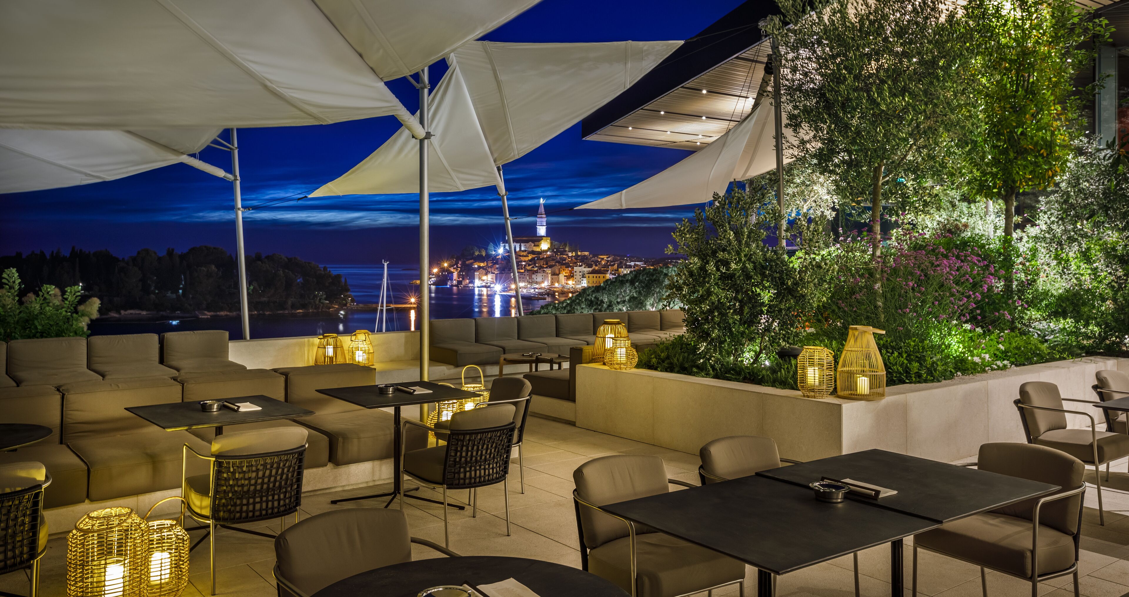 An outdoor patio with seating and tables, a view of a cityscape at night, and a balcony with plants and lighting.