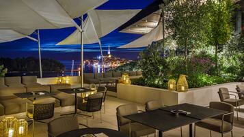An outdoor patio with seating and tables, a view of a cityscape at night, and a balcony with plants and lighting.