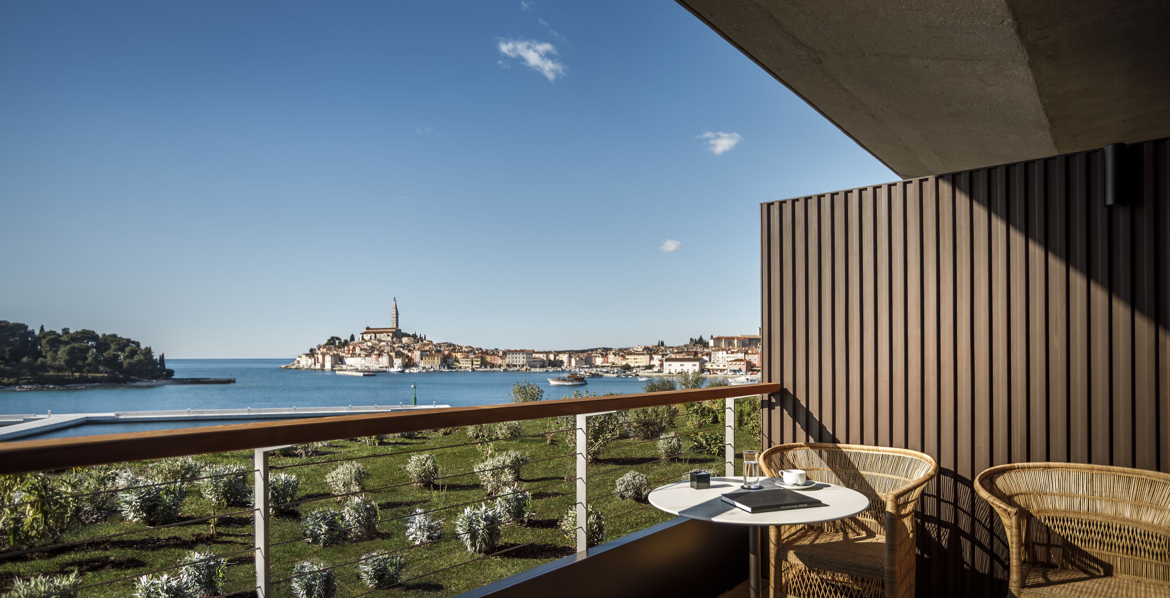 A balcony with a view of a coastal town, a table with chairs, and a garden.