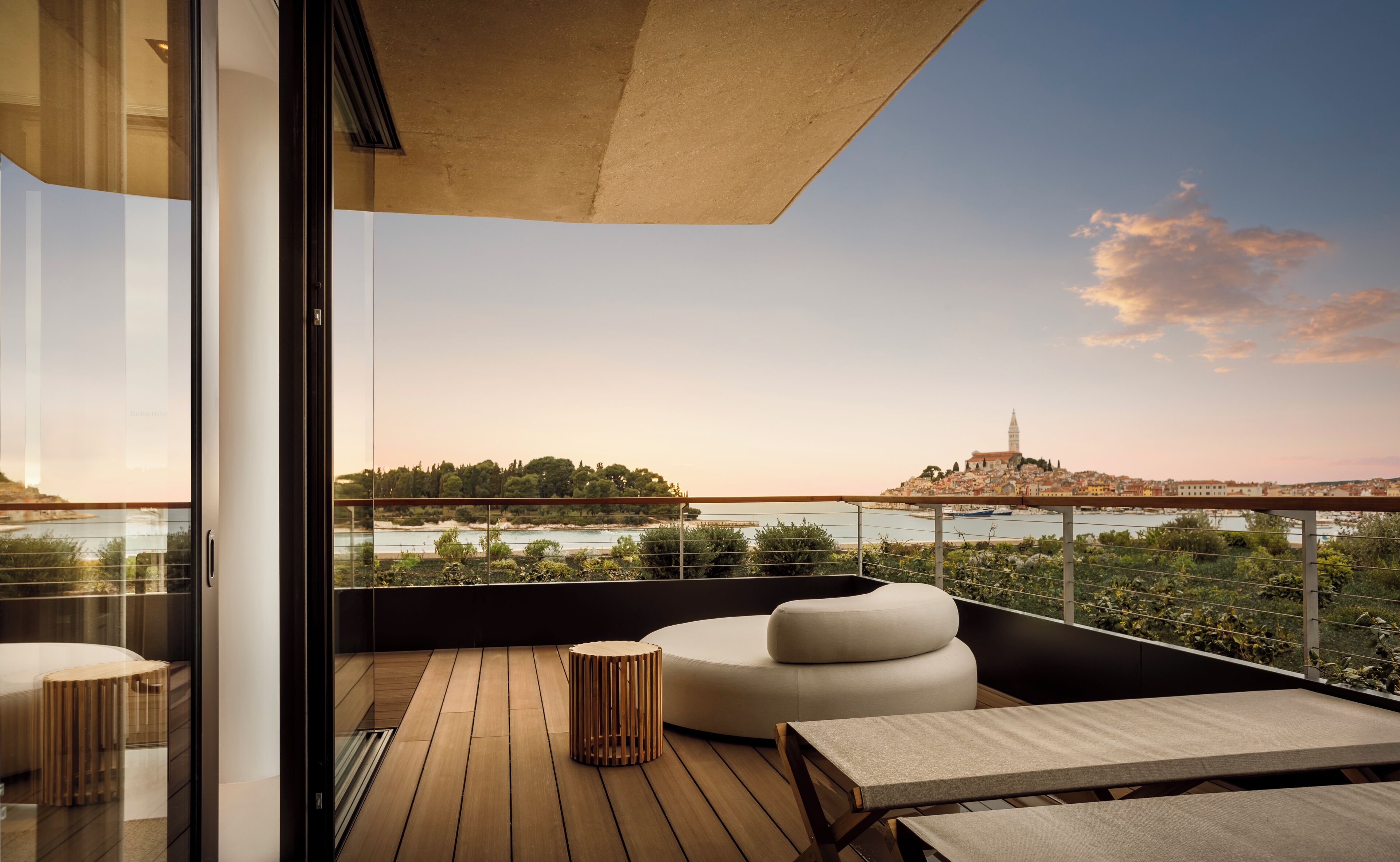 A modern balcony with wooden flooring, a round white sofa, and a small wooden table. A glass railing overlooks a scenic view of a city with a prominent tower.