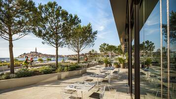 Een terras buiten met tafels en stoelen, met uitzicht op een kustplaats.