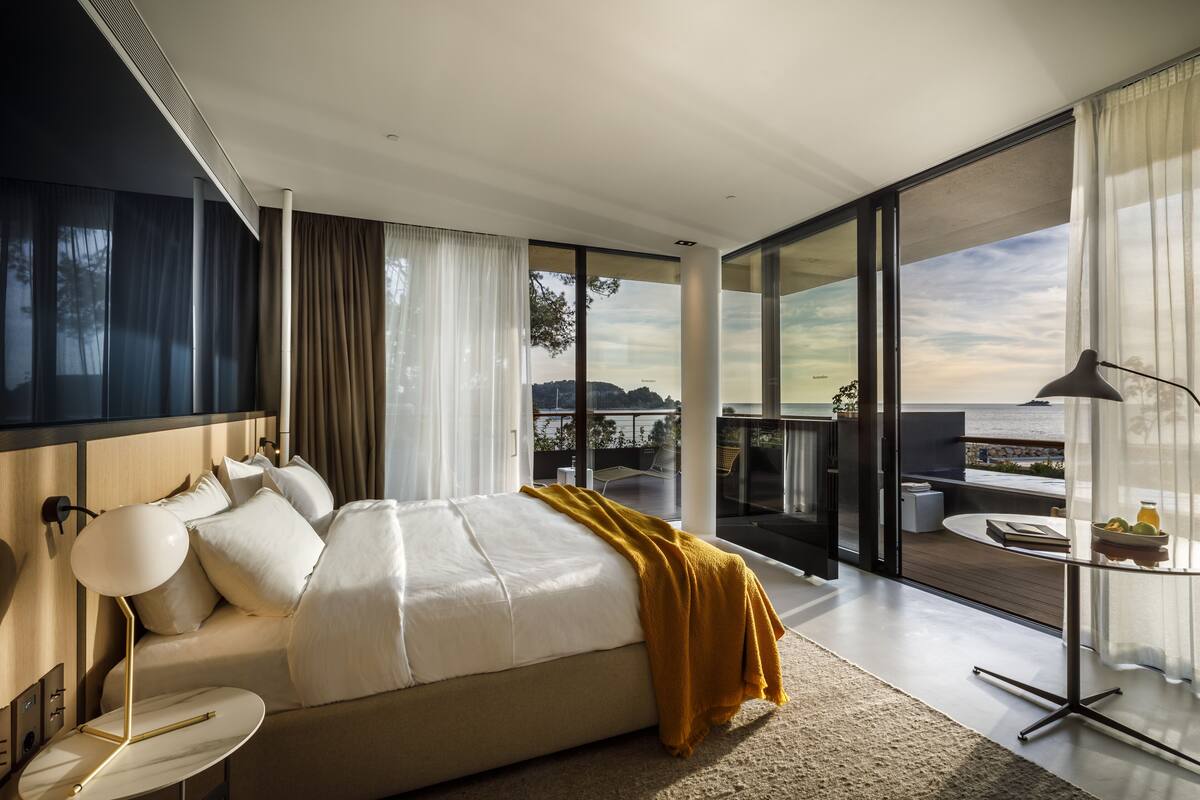 Un dormitorio moderno con una cama grande, una mesita de noche, una lámpara y vistas al exterior a través de amplias ventanas.