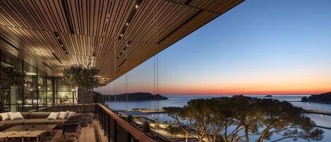 A modern outdoor lounge area with a wooden ceiling, seating, and a view of the sea at sunset.