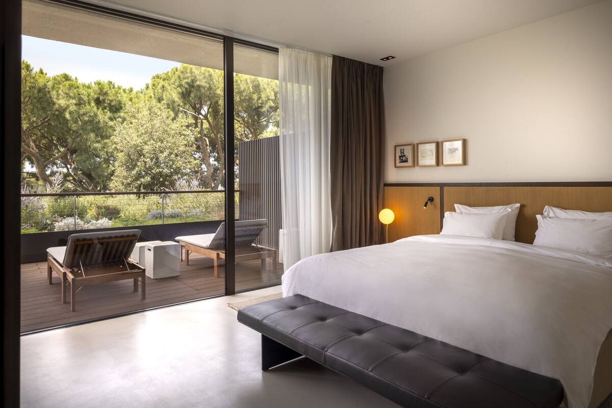 Habitación de hotel moderna con una cama grande, un banco, una mesita y vistas al exterior.