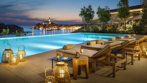 Ein Poolbereich mit Liegestühlen und Tischen, Blick auf eine Stadtlandschaft im Abendlicht und ein Gewässer.